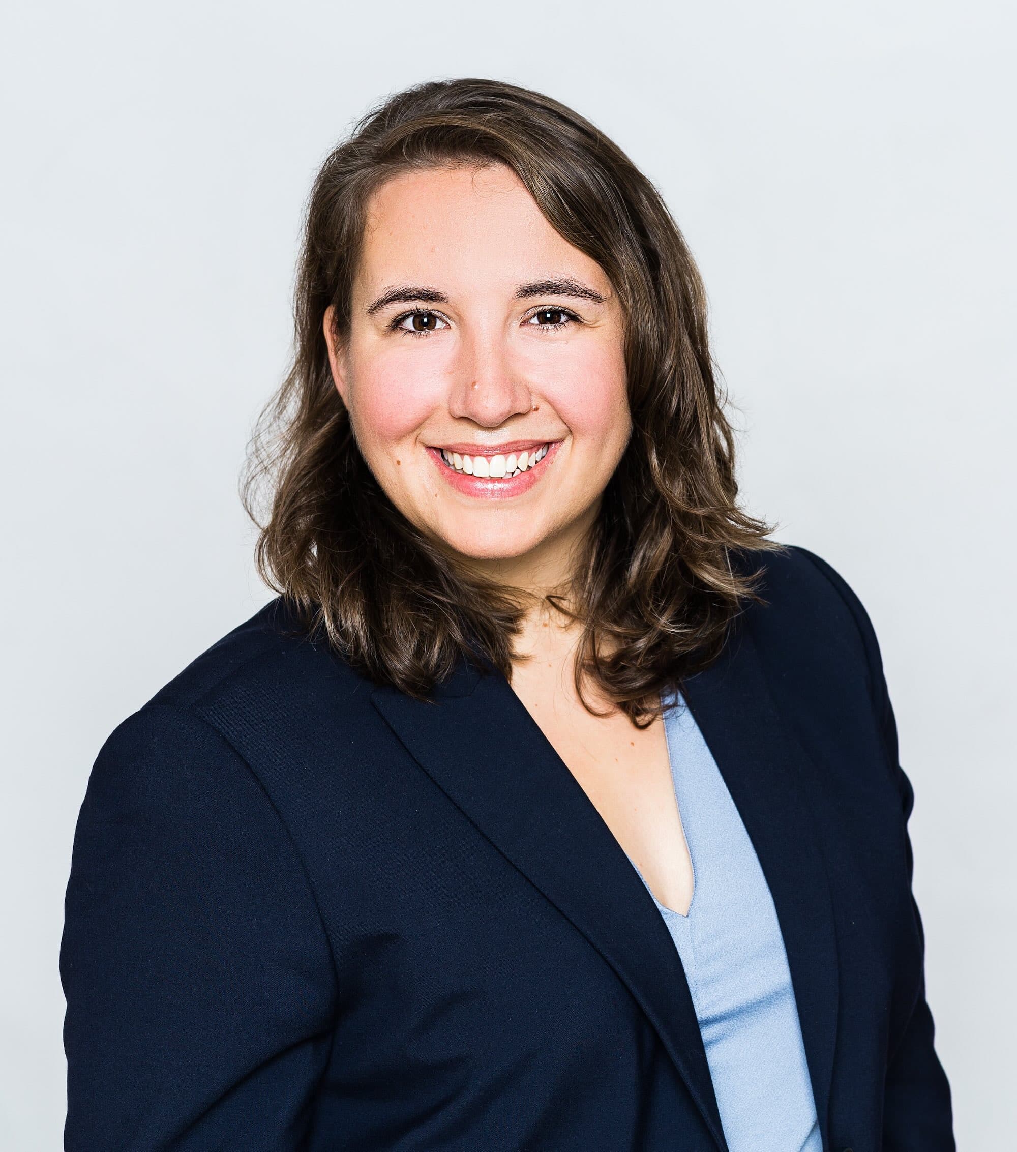 A woman with shoulder-length brown hair smiles at the camera and wears a dark blue blazer against a light background.