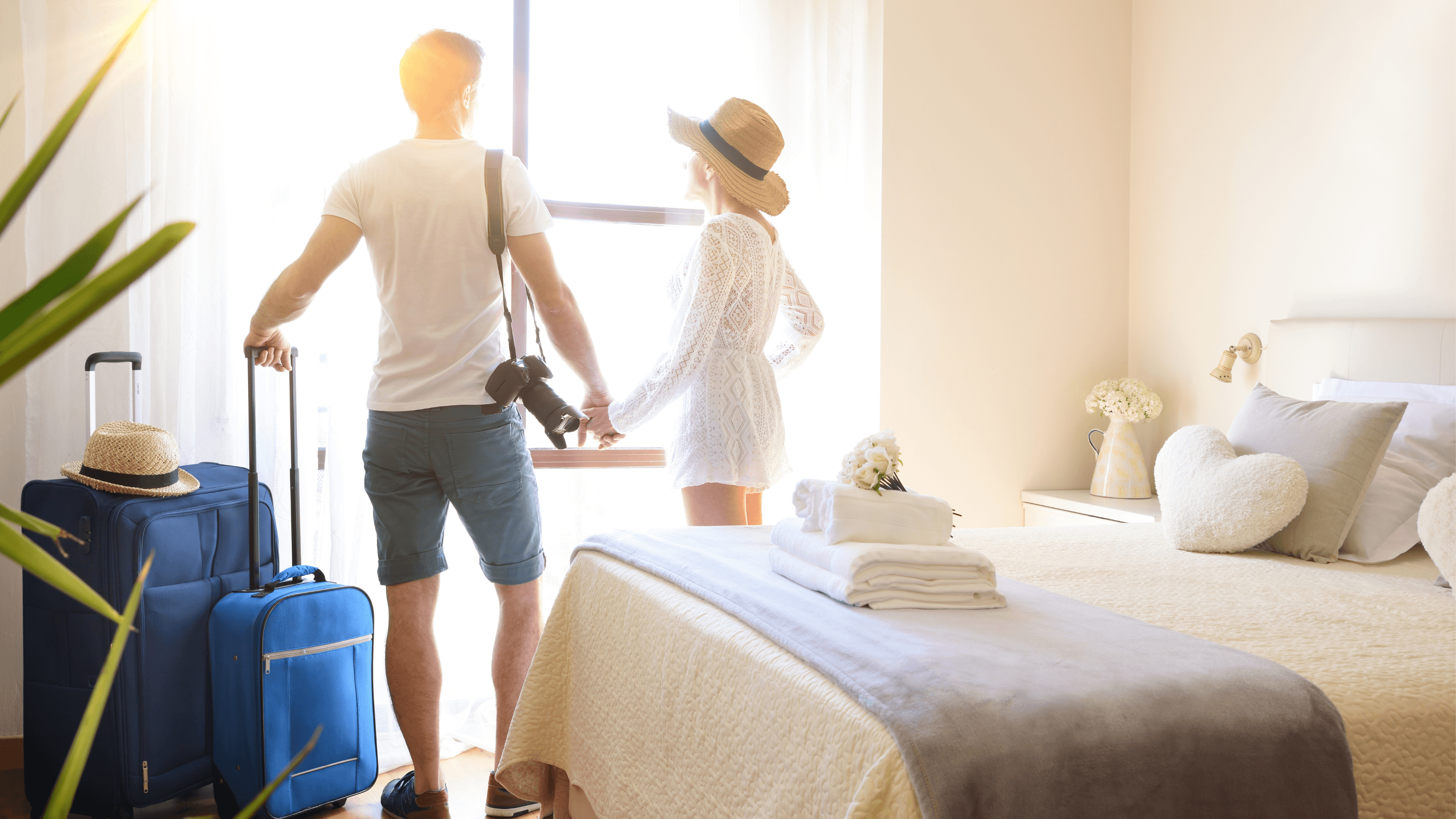 A couple stands with suitcases and a camera in a sunny hotel room and looks out the window.