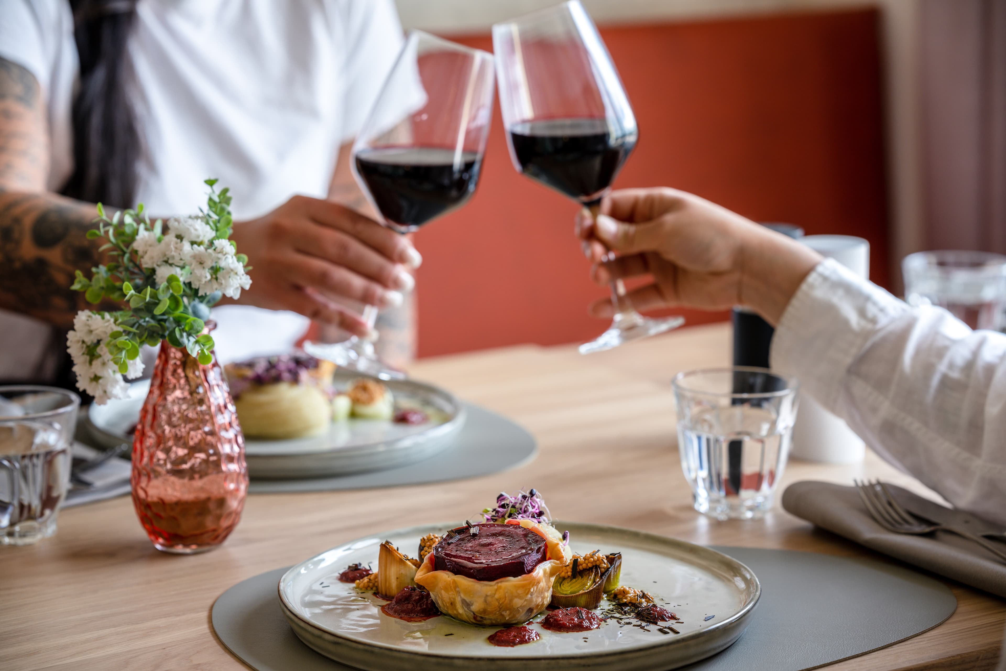 Two people clink red wine glasses at a table in a restaurant with food on a plate and a flower vase on it.