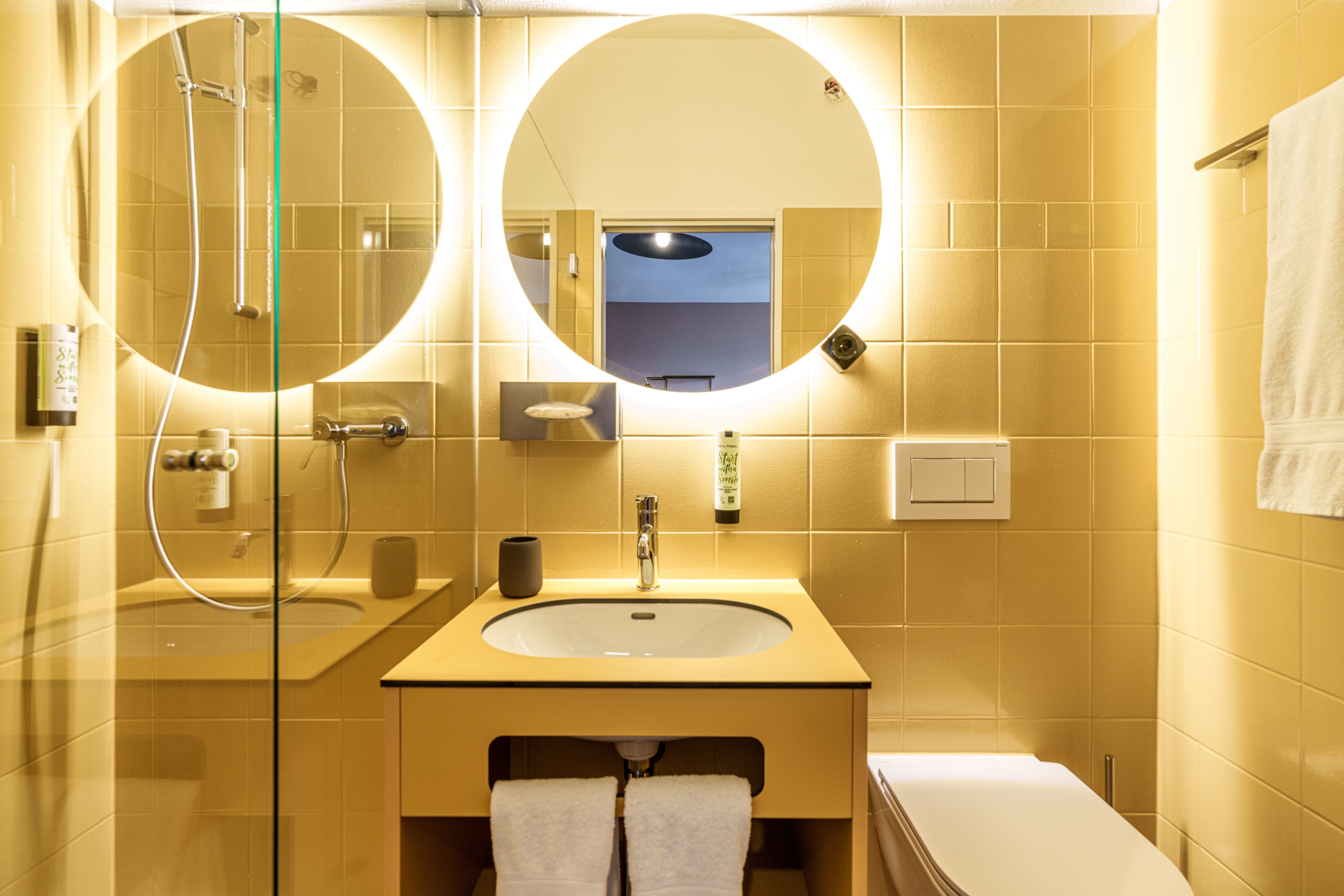 Modern bathroom in a double room at the B Smart Monastery with yellow tiles, an illuminated round mirror, a freestanding shower, and a sleek sink.